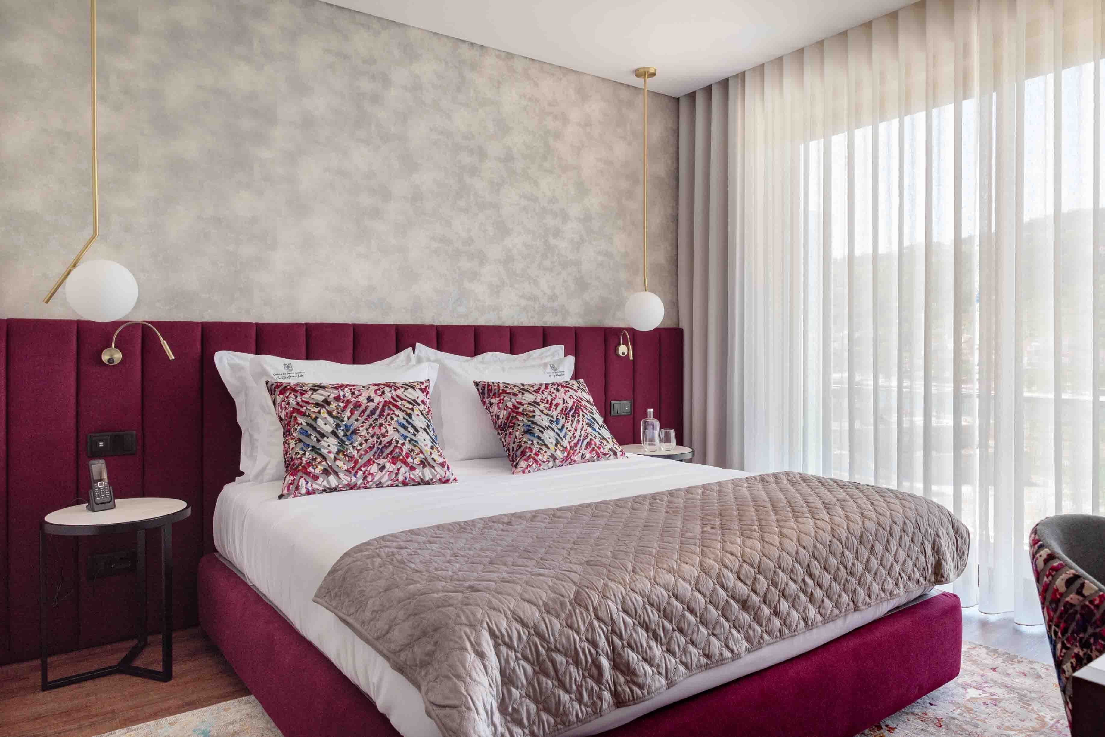 A modern hotel room with a large bed, a red upholstered headboard, a bedside table with a lamp, and a view of the outdoors through sheer curtains.