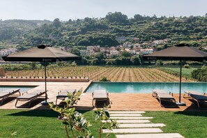 Zona de piscina con sillones y sombrillas, un viñedo al fondo y una colina con edificios.