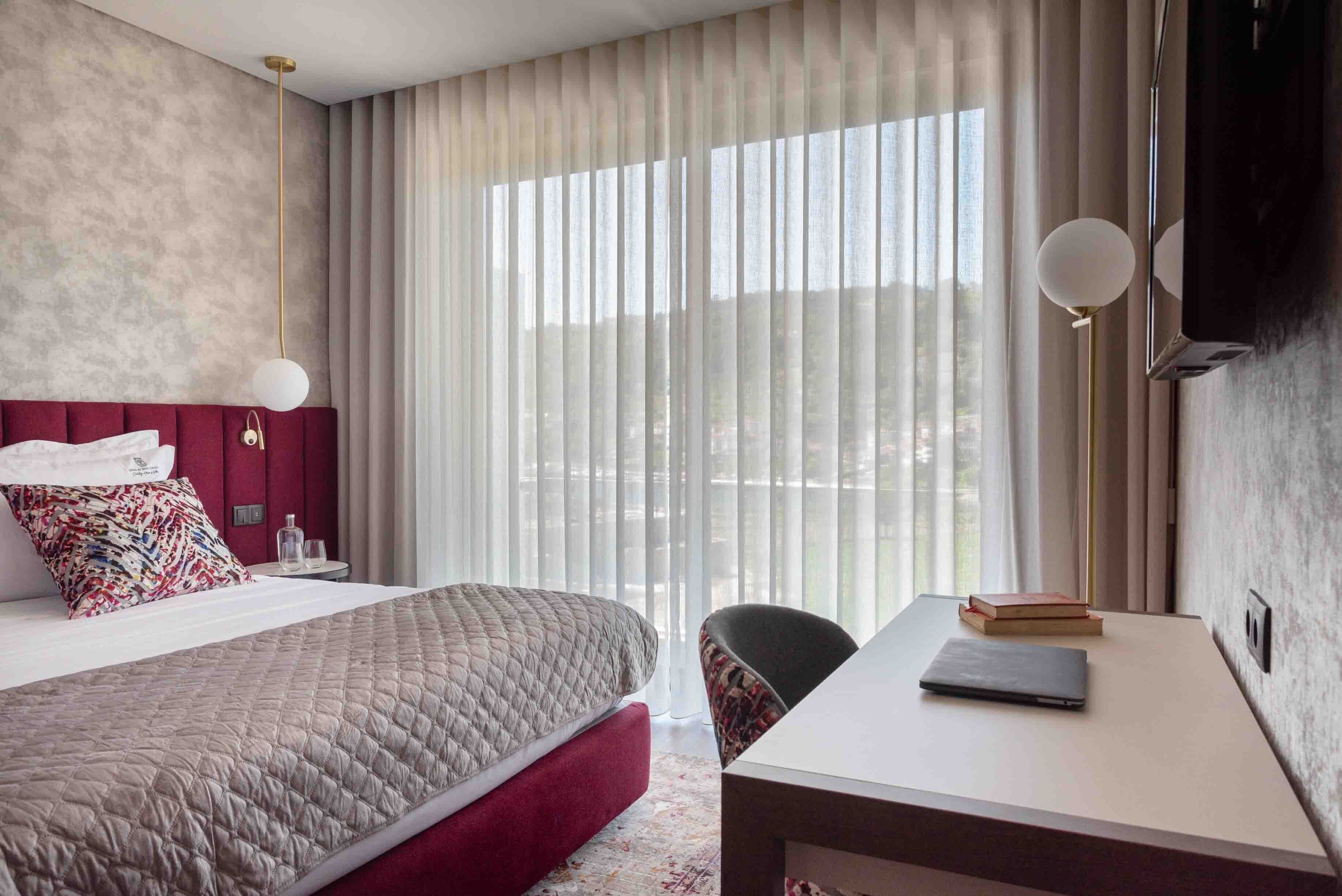 Habitación de hotel con cama, escritorio, silla, televisión y ventana con cortinas.
