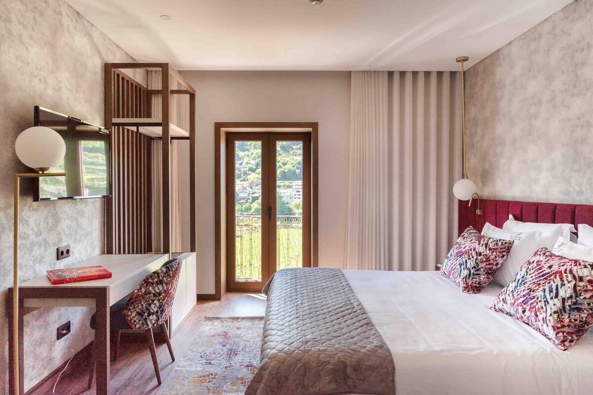 A modern hotel room with a large bed, a desk, and a view of the outdoors through a sliding door.