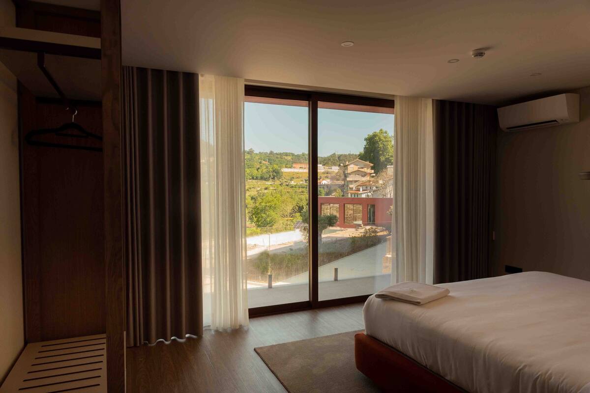 A hotel room with a large bed, a wardrobe, and a view of the city through floor-to-ceiling windows.
