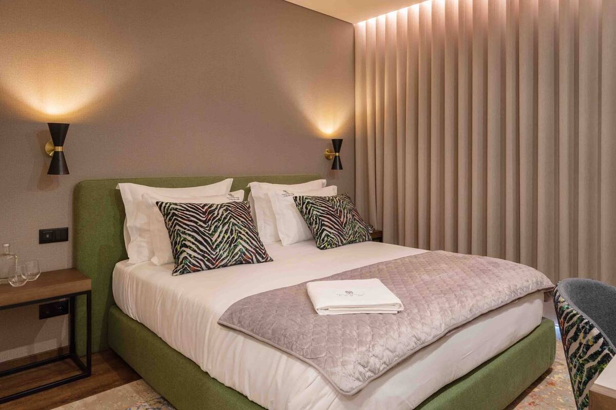 A neatly made bed with a green headboard, two patterned pillows, a folded white towel, and a bedside table with glasses.