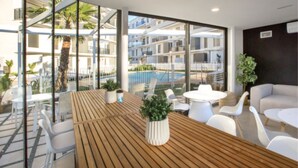 A modern hotel room with large windows, a wooden table, white chairs, and a view of a pool and buildings.