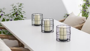 A white table with three candle holders, wicker chairs, and a white wall with a plant.