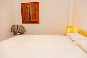 A bedroom with a wooden bed, a wicker chair, a window with wooden shutters, and a bedside lamp.