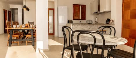 A dining area with a round table and chairs, a kitchen in the background, and a wooden panel on the right.