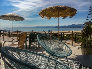 Une terrasse au bord de la plage, avec des chaises en osier, une table et deux parasols à toits de chaume.