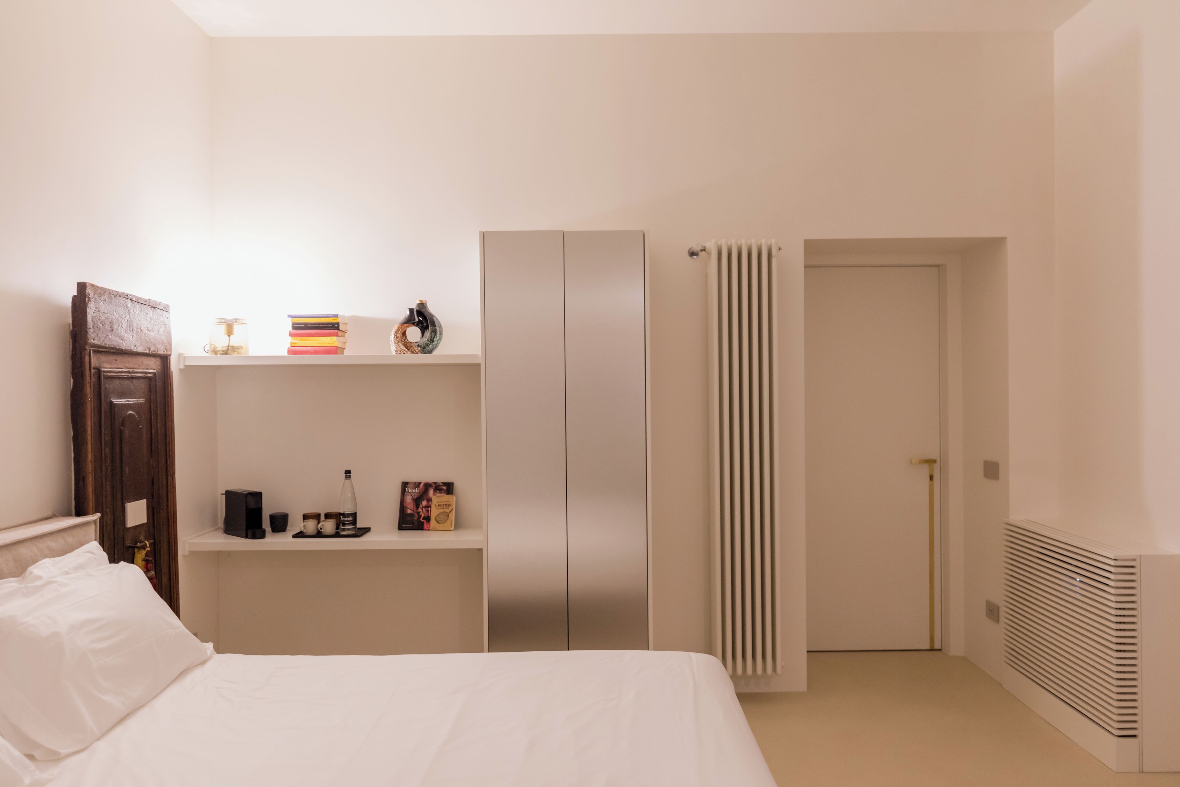 A bedroom with a bed, a wardrobe, a shelf with books and a bottle, and a radiator.