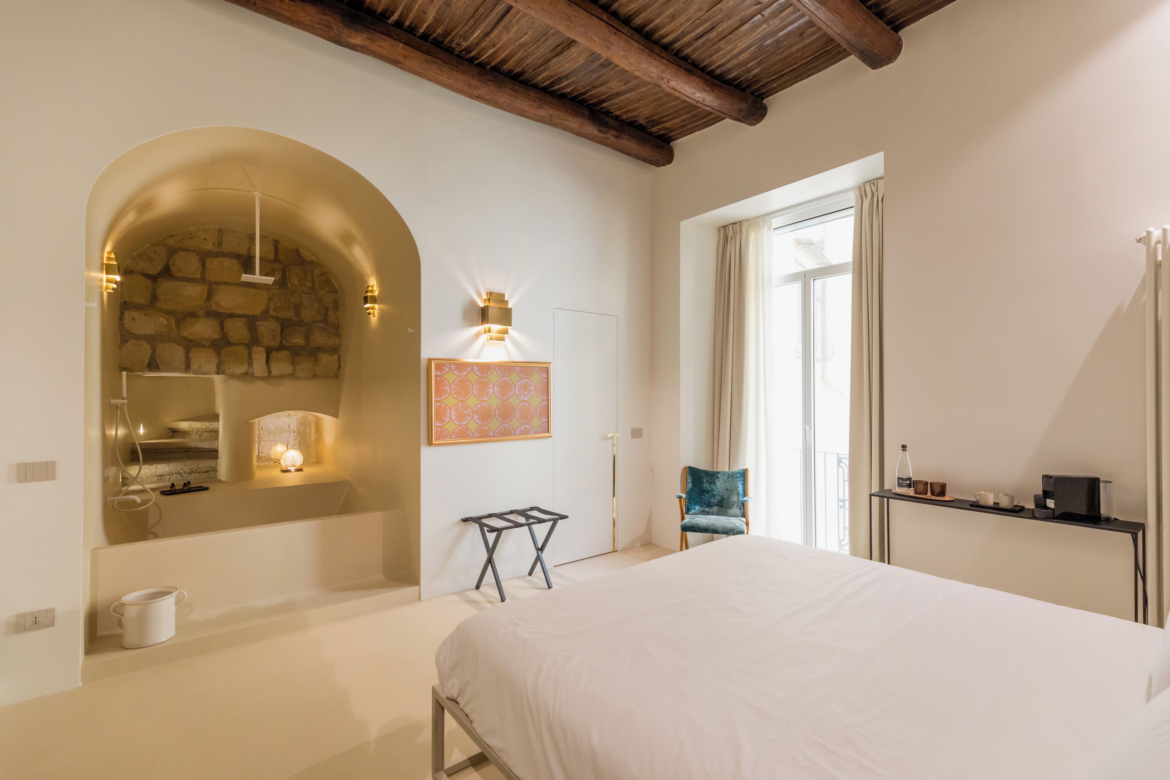A bedroom with a large bed, a stone wall arch, a small table, a chair, and a window with curtains.