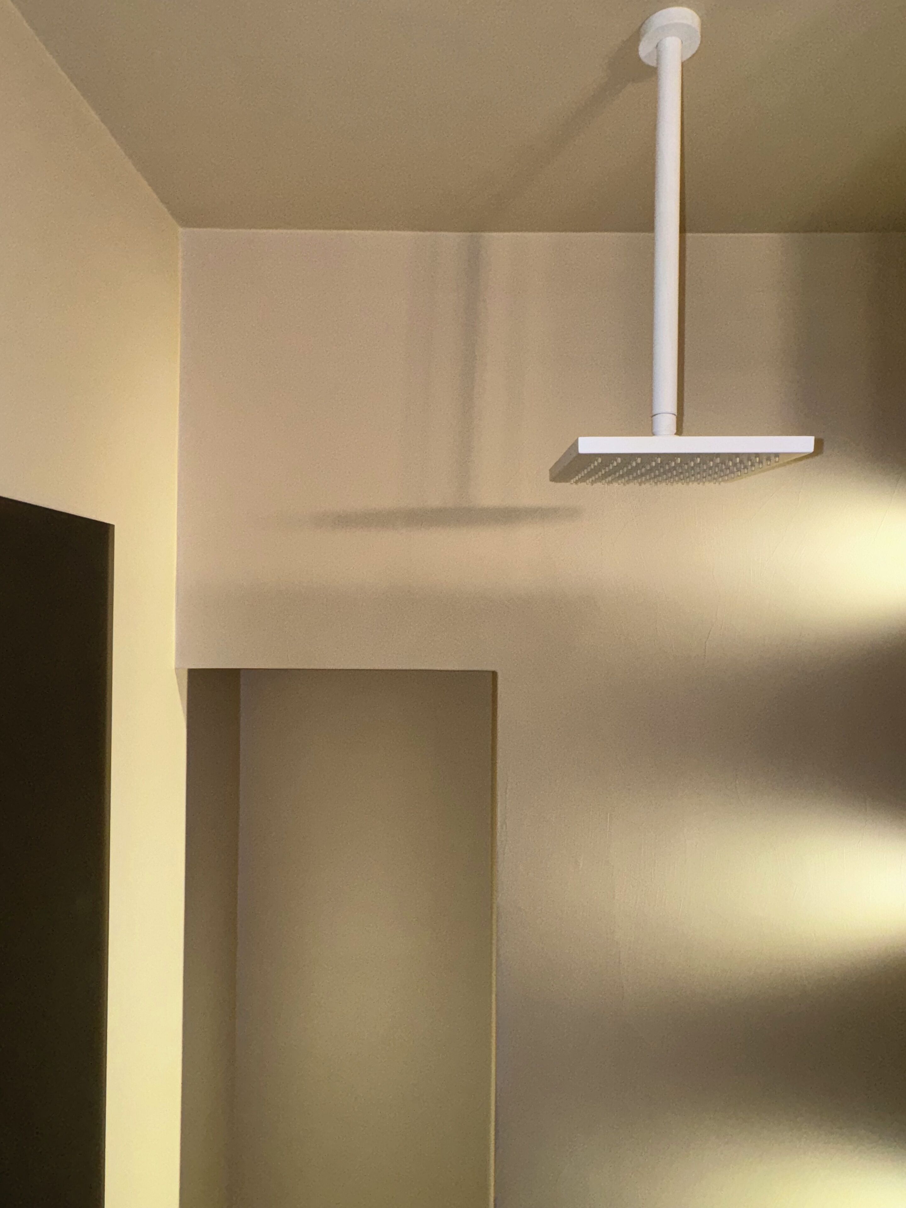 Casa de banho com chuveiro fixado numa parede branca, uma porta à esquerda e um candeeiro vertical.