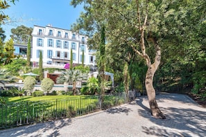 A building with multiple balconies, a paved walkway, and a well-maintained garden with a tree and a fence. - Château Les Alouettes (Cannes)