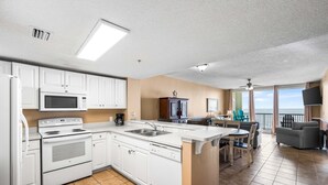 Cocina moderna con armarios blancos, horno empotrado y microondas. Cuenta con comedor con mesa y sillas, y balcón con vista al océano.