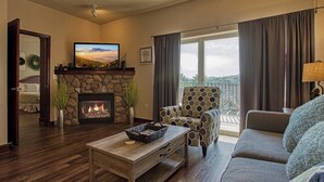 Ein Wohnzimmer mit Kamin aus Stein, einem darüber montierten Fernseher, einer Couch, einem Couchtisch und Blick auf einen Balkon mit Geländer.