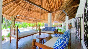 A covered outdoor seating area with a thatched roof, wooden furniture, and a view of the surrounding greenery. - Mfalme Luxury Villas (Mtwapa)