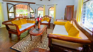 A room with wooden furniture, including sofas and chairs, a table with a vase, and a ceiling fan. - Mfalme Luxury Villas (Mtwapa)