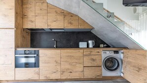 A compact kitchen with wooden cabinetry, a built-in oven, a washing machine, and a glass staircase. - Apartment In The Forest (Palanga)