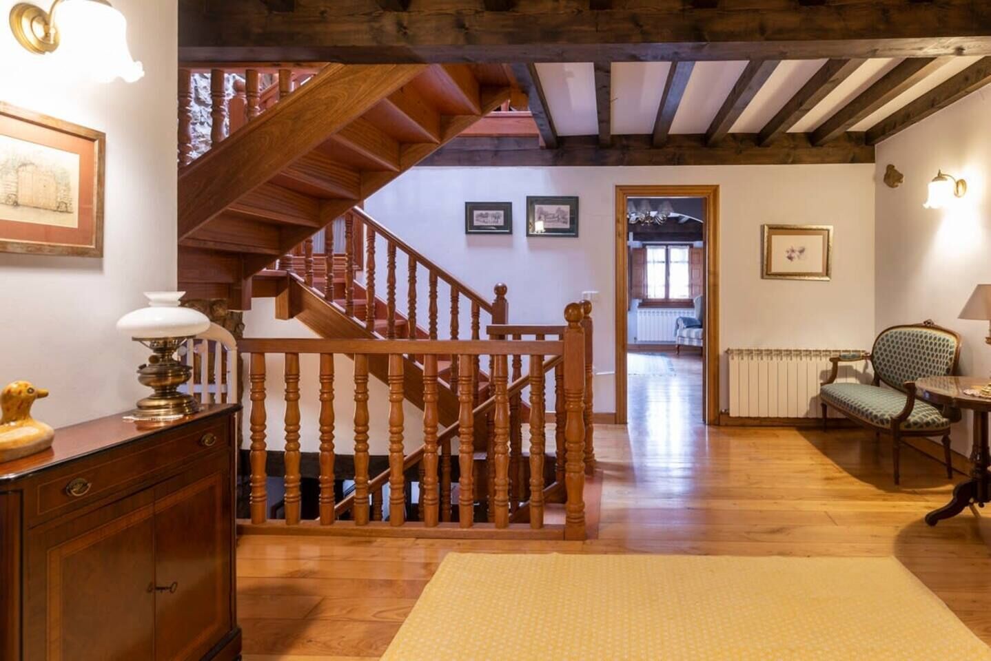 A wooden staircase with a railing leading to an upper floor, a patterned rug on the floor, and a chair with a lamp.
