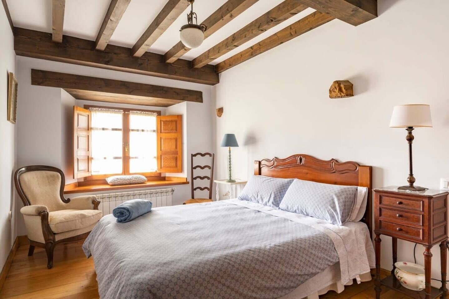 Un dormitorio con cama de madera, una silla, una mesita de noche, una lámpara y una ventana con contraventanas de madera.