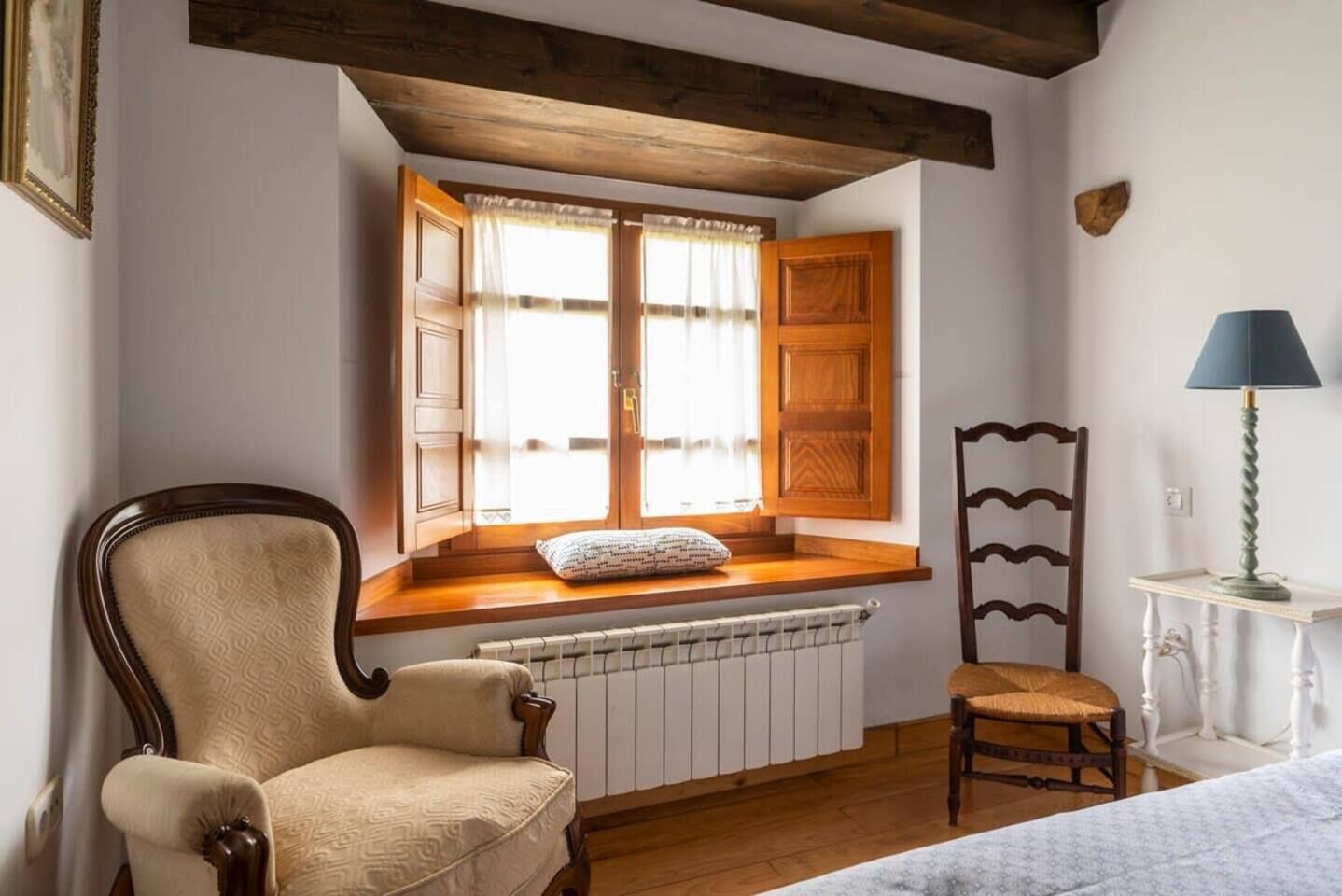 Una habitación con asiento junto a la ventana, un radiador, una silla, una lámpara y una butaca de madera.