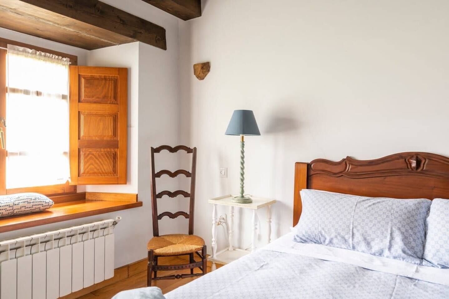Een slaapkamer met een houten hoofdbord, een nachtkastje met een lamp, een stoel en een raam met houten luiken.