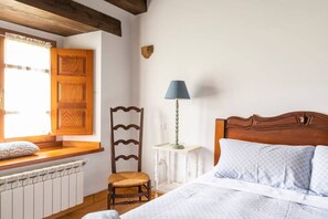 Un dormitorio con cabecera de madera, una mesita de noche con lámpara, una silla y una ventana con contraventanas de madera.