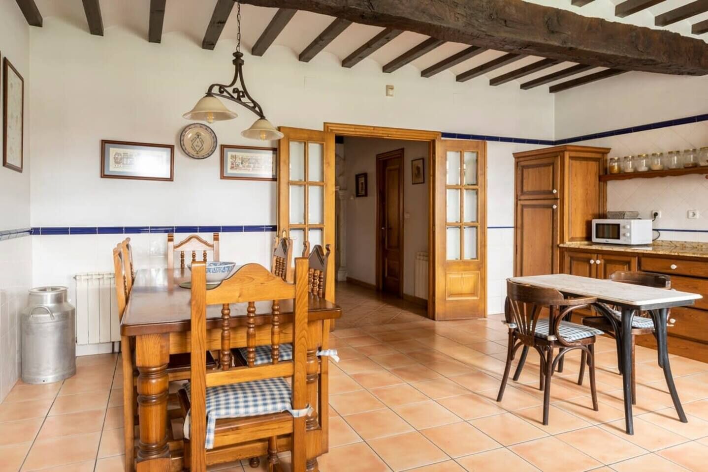 Une cuisine avec des meubles de rangement en bois, une table à manger, des chaises, un luminaire suspendu et un sol carrelé.