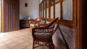 Une pièce avec un mur en briques, des poutres en bois au plafond et une grande fenêtre avec des rideaux. On y trouve des chaises en osier et une table sur laquelle est posé un saladier.