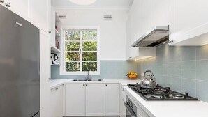 A modern kitchen with white cabinets, a stainless steel refrigerator, a gas stove, and a window with a view of greenery. - Charming Apartment | Malvern East (Malvern East)