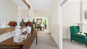 A hallway with a wooden dresser, a mirror, and a green armchair. There is a dining area in the background with a table and chairs. - Charming Apartment | Malvern East (Malvern East)
