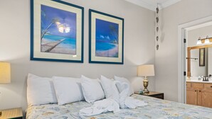 A hotel room with a bed, two framed beach paintings, a nightstand with a lamp, and a wooden cabinet with a mirror.