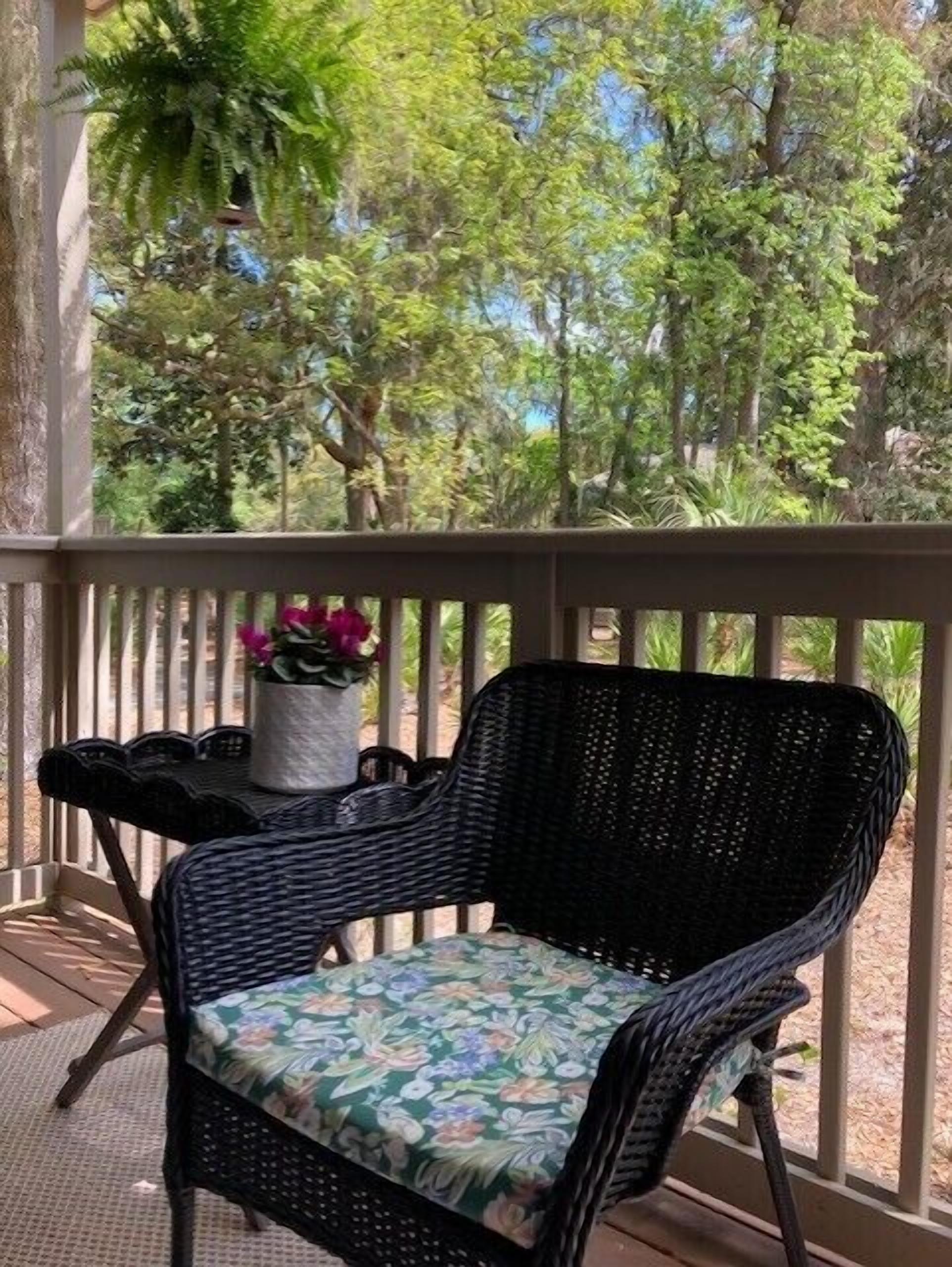 A wicker chair with a floral cushion on a wooden deck, with a potted plant and a table nearby.