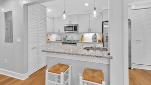 Cocina moderna con armarios blancos, encimeras de granito, microondas y refrigerador. Hay dos taburetes blancos con asientos de mimbre.