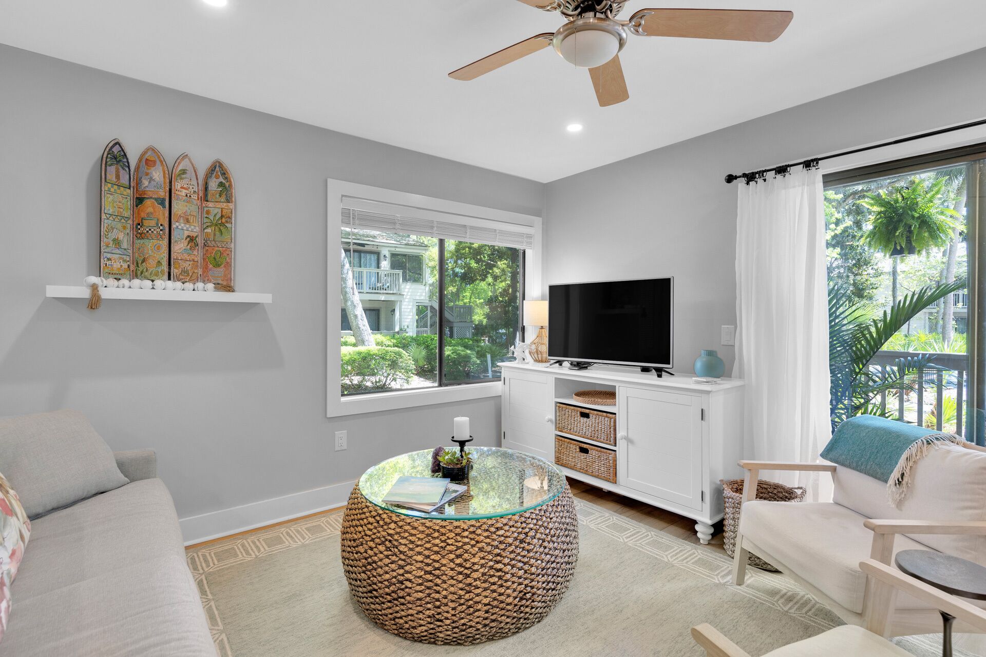 Un salon moderne avec un canapé, une table basse ronde en fibres tressées, un téléviseur posé sur un meuble blanc et un ventilateur de plafond.