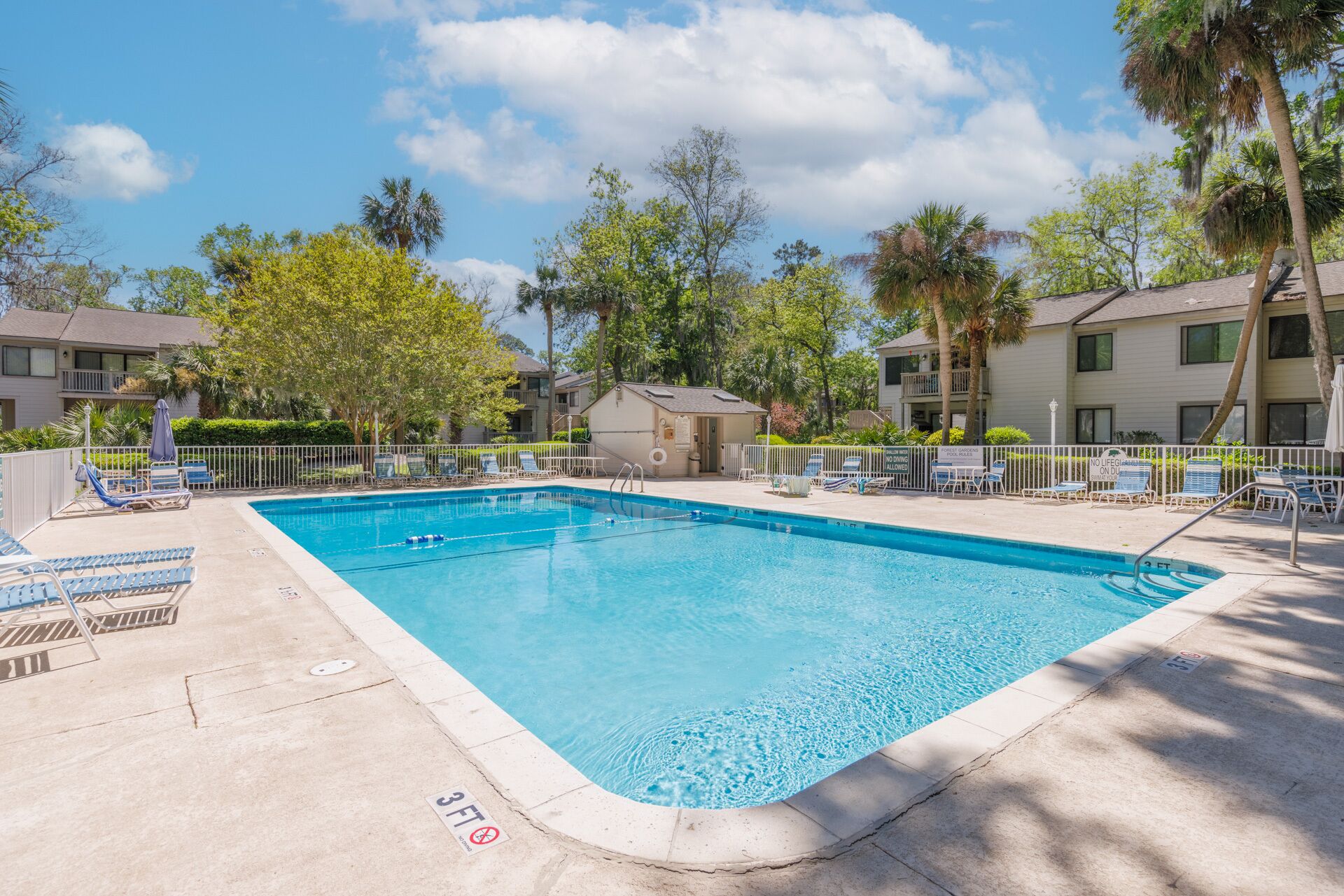 A rectangular swimming pool with a 3 FT depth marker, surrounded by lounge chairs and a small building, likely a changing room or storage, with a fence and trees in the background.
