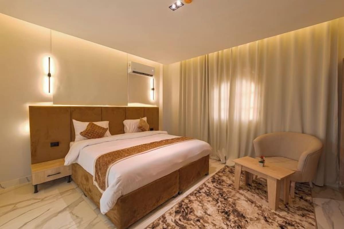 A hotel room with a large bed, a bedside table, a chair, a wall-mounted air conditioner, and a window with curtains.