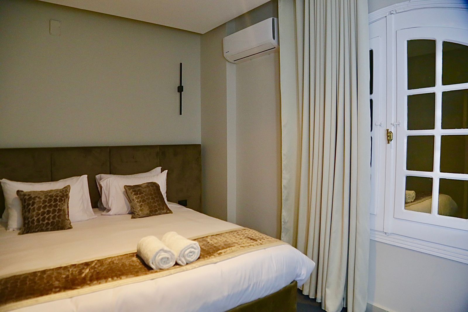 A hotel room with a large bed, two pillows, a headboard, a window with curtains, and a wall-mounted air conditioner.