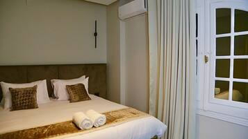 A hotel room with a large bed, two pillows, a headboard, a window with curtains, and a wall-mounted air conditioner.