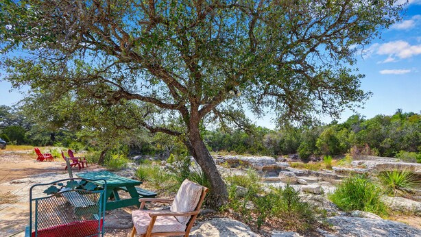 A picnic area with a table and chairs under a large tree, surrounded by rocks and greenery.