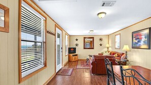 A cozy living room with a wooden floor, a brown leather sofa, a dining table with chairs, a TV, and a painting on the wall. - Hilltop Ranch Stay on 2,001 Acres | Shared Pool, Hot Tub & Trails (Johnson City)
