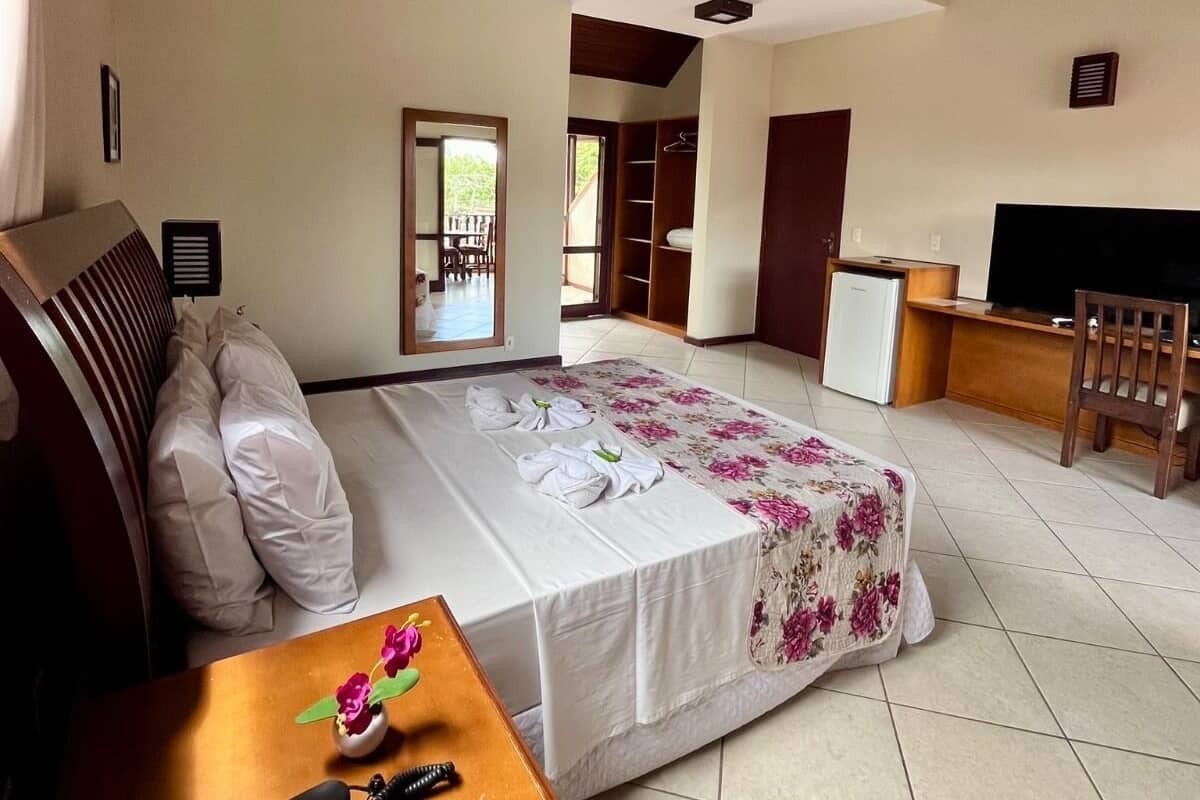 A hotel room with a bed, a desk, a chair, a small table with a remote control, and a window.