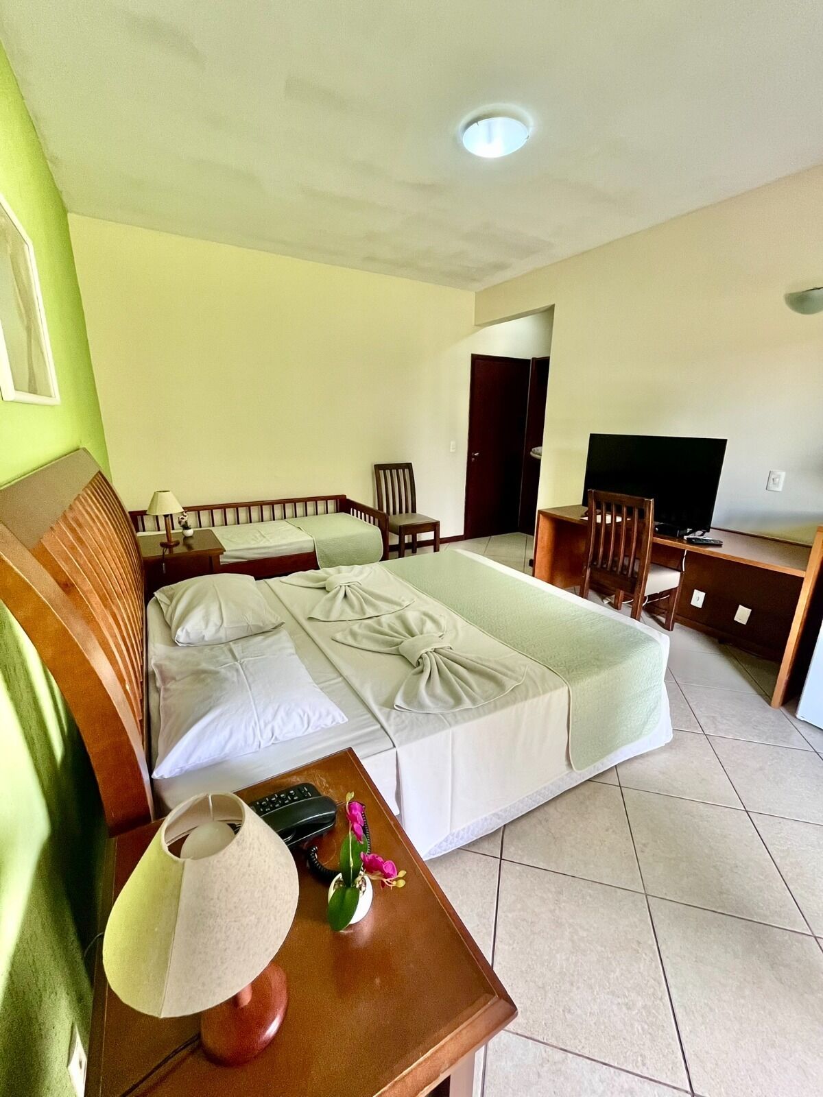 Una habitación de hotel con cama, mesita de noche, lámpara, controles remotos y televisor.