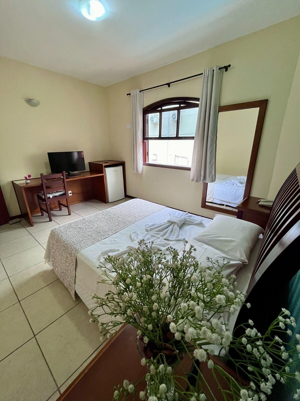 Habitación de hotel con cama, escritorio, televisor, refrigerador pequeño y ventana con cortinas.