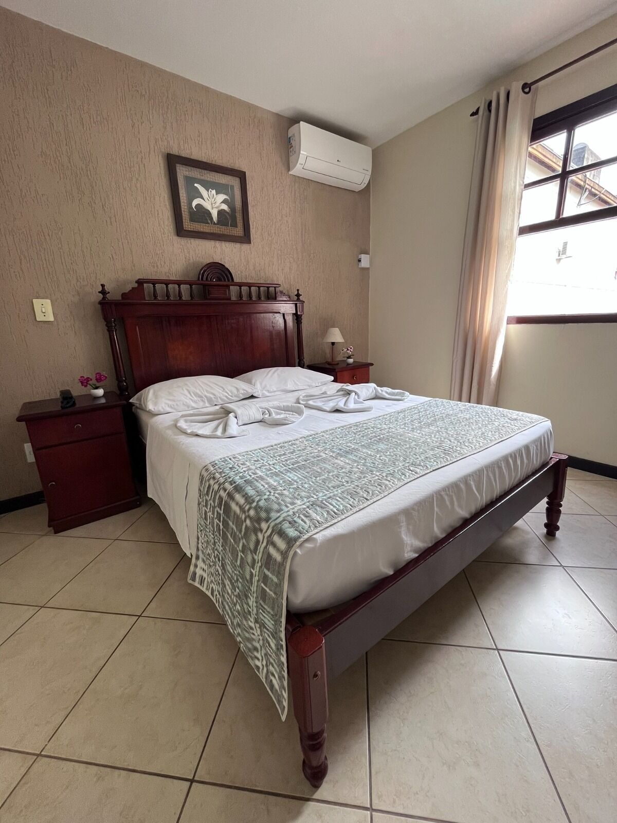 A neatly made bed with a patterned blanket, a wooden headboard, a nightstand with a lamp, and a wall-mounted air conditioner.