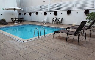 Una piscina rectangular con solárium empinado, rodeada de sillas de descanso y un muro blanco con plantas decorativas.