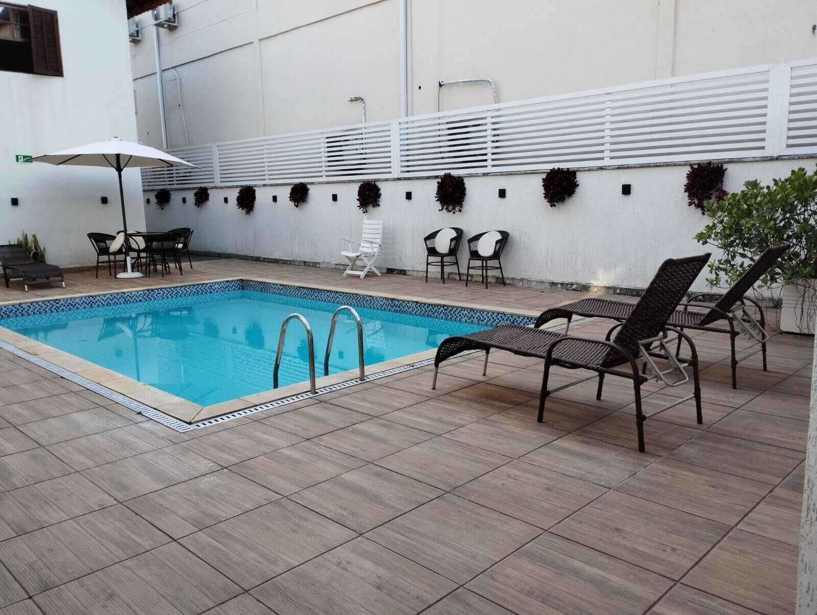 A swimming pool with tiled flooring, surrounded by lounge chairs and a white wall with decorative plants.