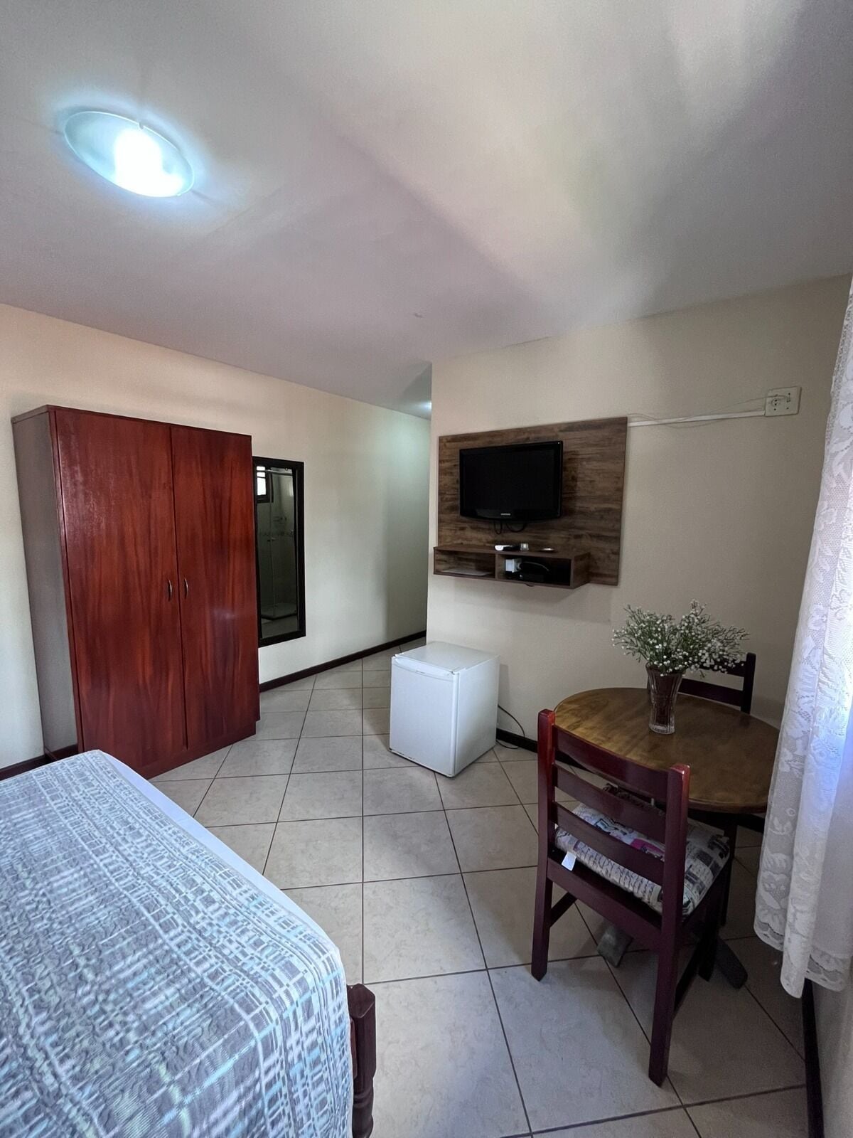A small, well-lit room with a bed, a wooden wardrobe, a television mounted on the wall, a small refrigerator, and a dining table with chairs.