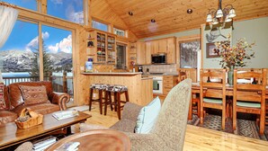 A spacious living area with a wooden ceiling, large windows offering mountain views, a kitchen with wooden cabinets, and a dining area with wooden chairs. - Lakeside Chalet - Luxury Retreat on Grand Lake w/ Dock & Kayaks (Grand Lake)