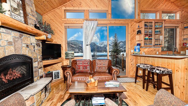 A cozy cabin interior with a stone fireplace, leather sofa, wooden dining table, and a kitchen area with a stone backsplash. - Lakeside Chalet - Luxury Retreat on Grand Lake w/ Dock & Kayaks (Grand Lake)