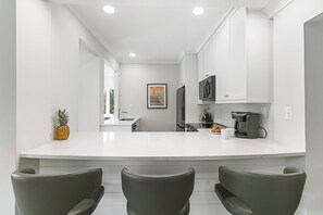 Une cuisine moderne avec des meubles blancs, un îlot central doté de sièges, une machine à café et un ananas posé sur le comptoir.