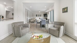 A modern living room with two grey sofas, a glass coffee table with a fruit bowl, a dining area with a white table and chairs, and a kitchen in the background. - Views and location, great villa in Turtle Point (Kiawah Island)